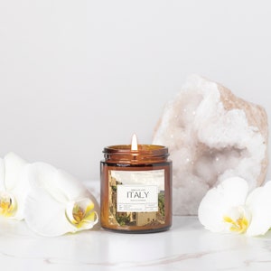 May include: A brown glass candle jar with a white label that says "Smells Like ITALY Fig & Cypress" and a picture of an Italian cityscape. The candle is lit and there are white orchids and a large white crystal on either side of the candle.