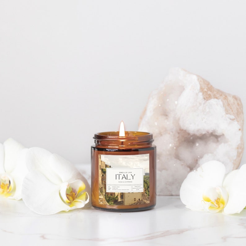 May include: A brown glass candle jar with a white label that says "Smells Like ITALY Fig & Cypress" and a picture of an Italian cityscape. The candle is lit and there are white orchids and a large white crystal on either side of the candle.