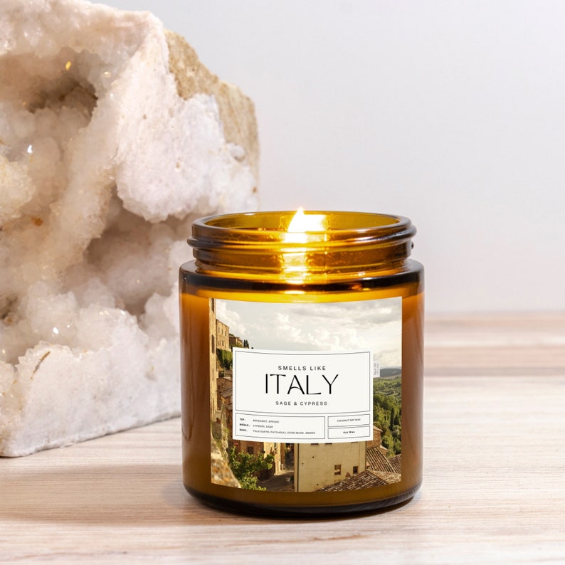 May include: A brown glass candle jar with a white label that reads "Smells Like Italy Sage & Cypress". The candle is lit and the jar is sitting on a wooden surface.