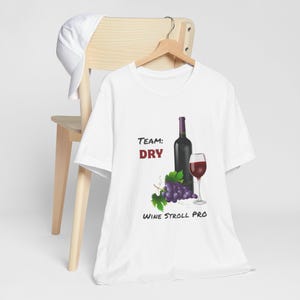 May include: White t-shirt with a wine-themed graphic. The design includes a wine bottle, a glass of red wine, grapes, and the text "Team: DRY" and "WINE STROLL PRO". The shirt is hanging on a wooden hanger.