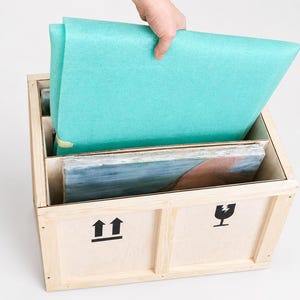Op de afbeelding: Een lichtgekleurde houten verzendkist met zwarte pijlen en symbolen voor breekbare goederen op de voorzijde. Binnenin de kist bevindt zich een rechthoekig voorwerp dat is ingepakt in een fel turquoise schuimvel en een ingelijst kunstwerk.