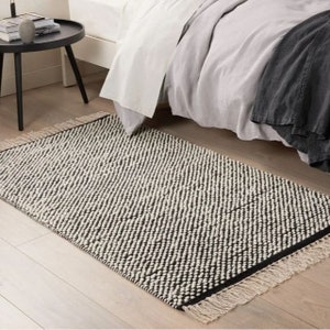 May include: Black and white patterned rug with a fringe border. The rug is made of a textured material and has a modern design.