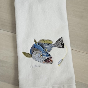 May include: White hand towel with an embroidered image of a speckled trout with a fishing lure. The embroidery is detailed and includes the signature "Carlton" in the bottom left corner.