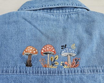 May include: A light blue denim shirt featuring an embroidered design of three mushrooms in orange, red, and blue, surrounded by small stitched leaves and stems. The embroidery is positioned horizontally across the back of the garment.