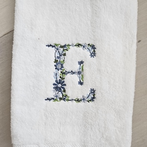 May include: A white terry cloth hand towel featuring an embroidered letter E. The letter is decorated with a detailed floral pattern in shades of blue, light blue, and green, with small leaves and blossoms forming the shape of the character.