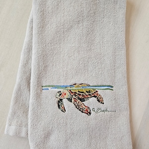 May include: A beige hand towel with an embroidered image of a sea turtle swimming underwater. The turtle is brown, green, and orange. The towel has a textured surface and a white border.