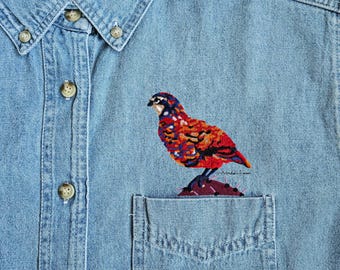 May include: A light blue denim button-down shirt featuring a colorful embroidered bird perched on a dark purple cactus. The bird has vibrant red, orange, and blue plumage. The shirt includes a chest pocket and a collar with buttons.