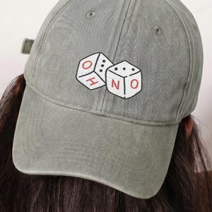 Puede incluir: Una gorra de béisbol color verde salvia con un bordado de dos dados blancos. Los dados están delineados en negro con letras rojas en las caras que forman la palabra OHNO. La gorra tiene una corona estructurada y una visera curva.