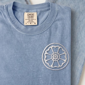 May include: A light blue t-shirt with a white embroidered circular lotus flower design on the chest and a Comfort Colors brand tag at the neck, paired with light wash blue jeans featuring a tan leather patch on the back waistband.