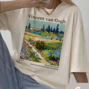May include: An ivory t-shirt featuring a graphic print of Vincent van Gogh's painting The Garden at Arles. The text reads Vincent van Gogh, (1889), The Garden at Arles, Kunstmuseum Den Haag, Netherlands. The shirt has short sleeves and a crew neck.