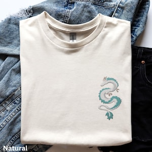 Puede incluir: Una camiseta de color crema con un gr&aacute;fico de drag&oacute;n verde y blanco en el lado izquierdo del pecho.