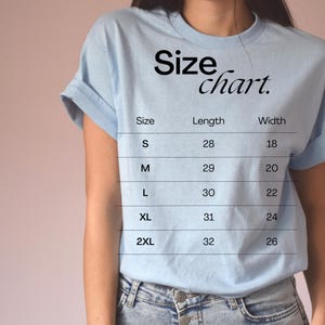 Puede incluir: Una camiseta azul claro con una tabla de tallas impresa en la parte delantera. La tabla muestra la talla, la longitud en pulgadas y el ancho en pulgadas para las tallas S, M, L, XL y 2XL.