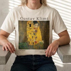 May include: A cream-colored short-sleeve t-shirt featuring a graphic print of Gustav Klimt's painting The Kiss. The text Gustav Klimt is printed above the artwork, with (1907-1908) and The Vienna on the bottom left and right corners respectively.