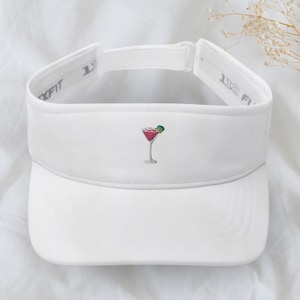 Puede incluir: Visera blanca con una pequeña imagen bordada de una copa de cóctel con una rodaja de lima. La visera tiene una correa ajustable negra con el texto "1.5" en la correa.