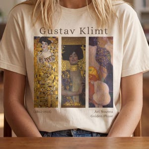 Può includere: Una t-shirt color crema con il testo Gustav Klimt in alto e (1862-1918) Art Nouvea Golden Phase in basso. Il centro mostra tre pannelli verticali con riproduzioni dettagliate di dipinti di Gustav Klimt.