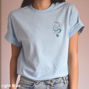 Puede incluir: Una camiseta azul claro con un dise&ntilde;o de drag&oacute;n verde y blanco en la parte delantera.