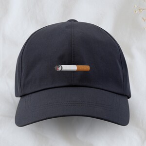 Könnte beinhalten: Eine dunkelblaue Baseballkappe mit einem weißen Zigaretten-Motiv auf der Vorderseite.