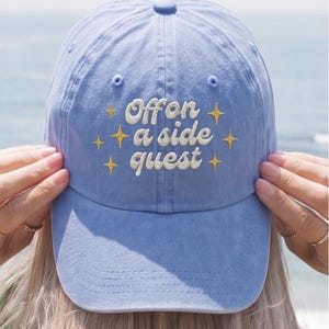 Könnte beinhalten: Eine hellblaue Baseballkappe mit dem gestickten weißen Text "Off on a side quest" in Schreibschrift, umgeben von sechs kleinen, vierzackigen gelben Sternsymbolen. Die Kappe hat eine strukturierte Krone, einen gebogenen Schirm und einen Knopf.