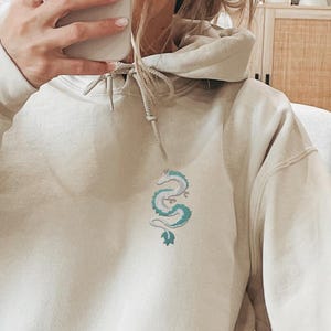 Könnte beinhalten: Ein cremefarbener Hoodie mit einem blauen und weißen Drachen, der auf der Vorderseite gestickt ist.