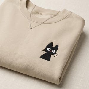 Könnte beinhalten: Ein beigefarbenes Sweatshirt mit Rundhalsausschnitt und einer kleinen Stickerei eines schwarzen Katers mit großen weißen Augen und einem winzigen roten Mund auf der Brust. Eine dünne silberfarbene Kette liegt um den Halsausschnitt des langärmeligen Kleidungsstücks.