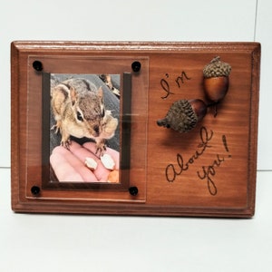 May include: A wooden picture frame with a photo of a chipmunk eating peanuts. The frame has a clear acrylic insert and two acorns attached to the right side. The text "I'm about you!" is engraved on the wood.