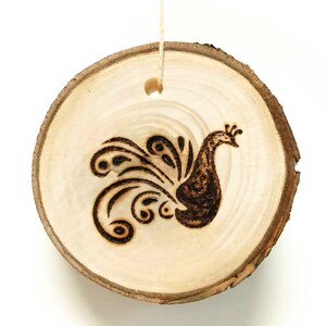Pode incluir: Um ornamento de madeira em forma de fatia de madeira com um design de pavão queimado em madeira, com a cauda espalhada.