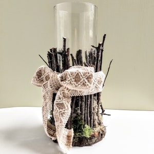 May include: A glass cylinder vase filled with twigs and moss, wrapped with a beige burlap ribbon. The vase is sitting on a white surface.