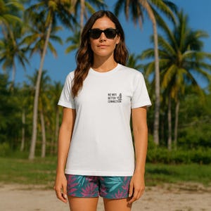 Op de afbeelding: Wit T-shirt met de tekst "NO WIFI BETTER CONNECTION" en een surfplankafbeelding. De persoon draagt een zwarte zonnebril en een kleurrijke korte broek met een tropisch patroon. Palmbomen en een blauwe lucht op de achtergrond.