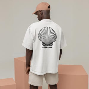 May include: White t-shirt featuring a black and white seashell design on the back, with the word "SURFSURF" underneath. The person is wearing a beige baseball cap and beige shorts. The shirt has a loose fit.