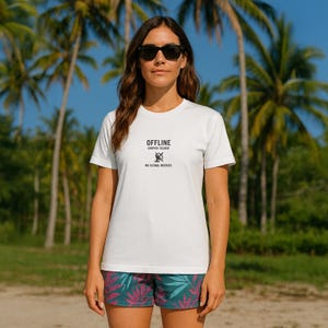 Op de afbeelding: Een wit T-shirt met de zwarte tekst "OFFLINE SURFER ISSUED NO SIGNAL NEEDED". Het shirt wordt gecombineerd met een short met teal- en roze patronen. Op de achtergrond palmbomen en een blauwe lucht.