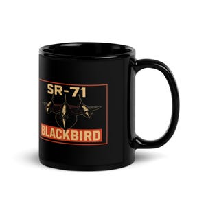 May include: Black ceramic mug featuring the text "SR-71 BLACKBIRD" in a rectangular orange and black design. The design includes an illustration of the SR-71 Blackbird aircraft. The mug has a black handle and a glossy finish.