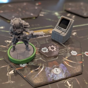 May include: A grey miniature soldier figure on a green base, alongside a small grey control panel, sits on a game board. The board features numbered sections and text, indicating a strategy game or war game setting.