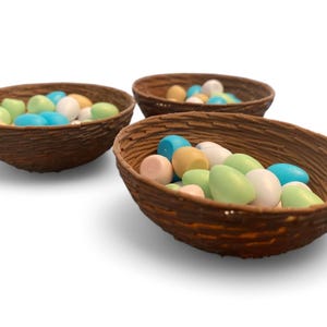 以下が含まれることがあります： 3つの茶色の編まれたバスケットに、パステルカラーのキャンディーの卵が入っています。卵は青、緑、ピンク、ベージュの色合いです。