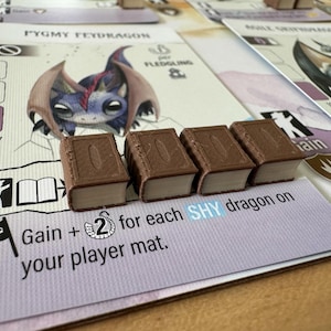 May include: A board game setup featuring cards with dragon illustrations and miniature book-shaped game pieces. The cards display text including "PYGMY FEYDRAGON" and "Gain + 2 for each SHY dragon on your player mat."