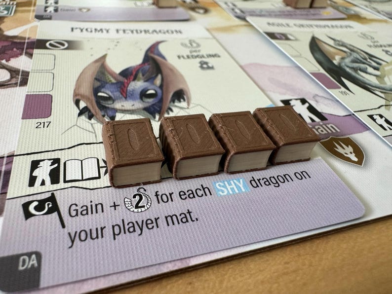 May include: A board game setup featuring cards with dragon illustrations and miniature book-shaped game pieces. The cards display text including "PYGMY FEYDRAGON" and "Gain + 2 for each SHY dragon on your player mat."