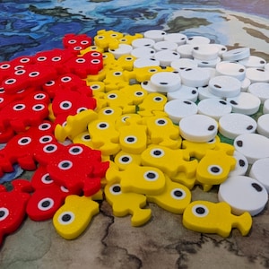 Op de afbeelding: Een verzameling kleurrijke spelstukken in rood, geel en wit. De rode stukken zijn onregelmatig van vorm met meerdere oogachtige cirkels. Gele stukken zijn visvormig met een enkel oog. Witte stukken zijn rond met een zwarte stip.