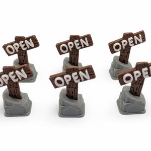 Könnte beinhalten: Sechs Miniatur-Holzschilder mit dem Wort "OPEN" in weißer Schrift. Die braunen Schilder sind auf grauen Steinsockeln montiert. Sie stehen vor einem weißen Hintergrund.