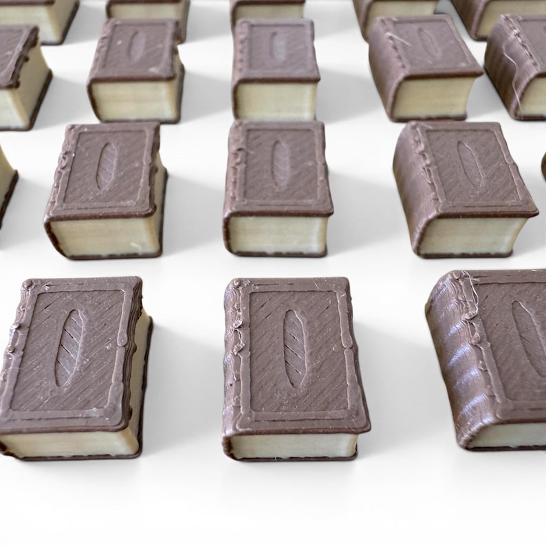 May include: Rows of chocolate candies shaped like miniature books. Each candy has a light-colored filling and a dark brown chocolate exterior, with a detailed book cover design. The candies are arranged on a white surface.