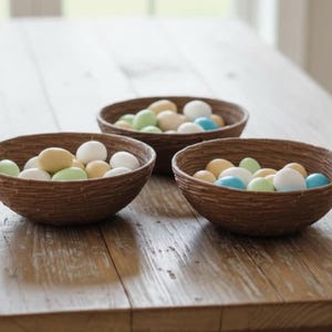 Puede incluir: Tres cuencos marrones tejidos llenos de caramelos de colores pastel, incluyendo amarillo, verde, azul y blanco, están sobre una mesa de madera. Los cuencos tienen una apariencia texturizada y rústica, y los caramelos tienen forma de huevo.