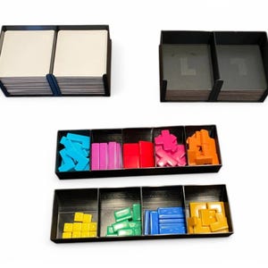 May include: Black game organisers with various coloured game pieces and cards. The organisers are divided into compartments, holding blue, pink, red, orange, yellow, and green game components. The cards are stacked in two separate organisers.