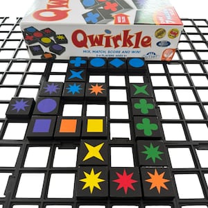 Puede incluir: Un juego de mesa Qwirkle con fichas negras que presentan varias formas y estrellas de colores. La caja del juego es visible, mostrando el nombre y las instrucciones del juego. El juego está diseñado para 2-4 jugadores.