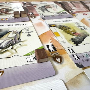 May include: A tabletop game with illustrated cards featuring dragons and other fantasy elements. The cards have text and icons, with small wooden game pieces scattered around. The game cards are white with colorful illustrations.