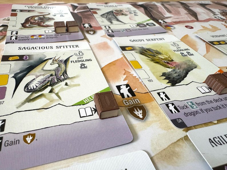 May include: A tabletop game with illustrated cards featuring dragons and other fantasy elements. The cards have text and icons, with small wooden game pieces scattered around. The game cards are white with colorful illustrations.