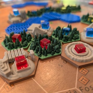 以下が含まれることがあります： 3Dプリントされた地形ピースを備えたボードゲーム。ゲームボードには、ミニチュアの森、構造物、水の特徴を備えた六角形のタイルがあります。赤と青の半透明の立方体が地形に配置されています。ゲームピースはグレー、グリーン、ブラウンの色合いです。