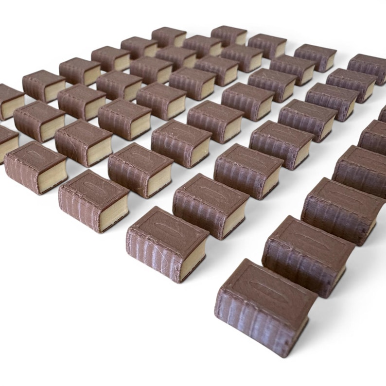 May include: Rows of miniature chocolate book-shaped candies with brown covers and tan-colored pages. The candies are arranged in neat rows on a white surface, creating a visually appealing pattern. The chocolate appears to have a textured surface.