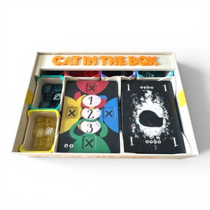 May include: An open board game box labelled "CAT IN THE BOX" containing game cards and pieces. The cards feature numbers and symbols, with small containers holding tokens. The game cards have the numbers 1, 2, and 3. The box is white with orange text.