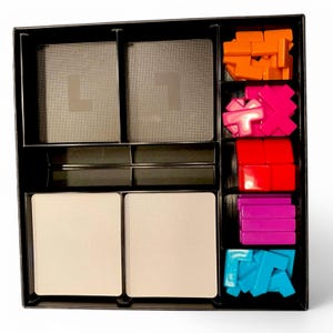 May include: A black game organiser with several compartments. The organiser contains cards with the letter "L", and various colourful game pieces in orange, pink, red, purple, and blue. The game pieces are different shapes.