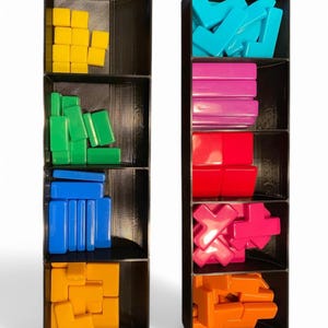 May include: Two black organisers, each with multiple compartments, filled with colourful game pieces. The pieces are various shapes and colours, including yellow squares, green rectangles, and blue bars. The other organiser contains teal L-shapes, pink rectangles, and red blocks.