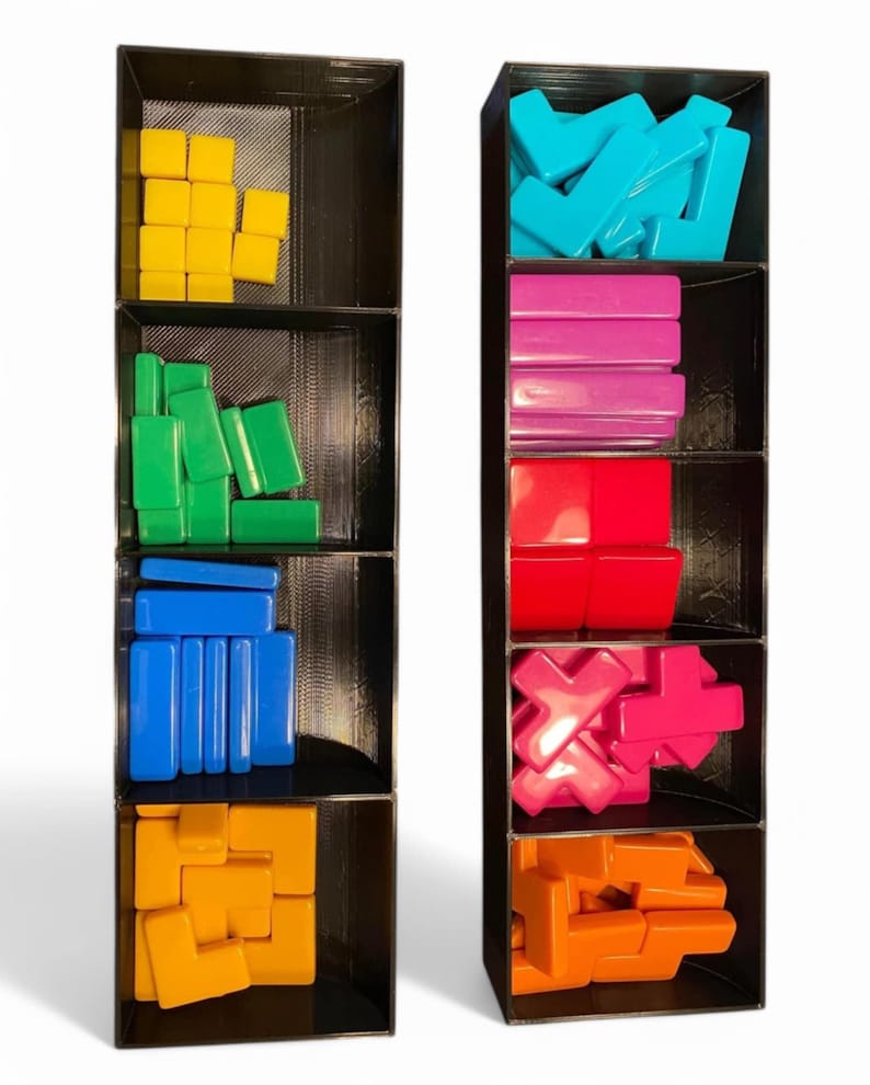 May include: Two black organisers, each with multiple compartments, filled with colourful game pieces. The pieces are various shapes and colours, including yellow squares, green rectangles, and blue bars. The other organiser contains teal L-shapes, pink rectangles, and red blocks.
