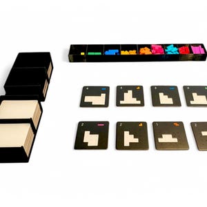 May include: A board game set with black components and colourful pieces. The set includes a stack of black boxes with white cards, a tray with coloured blocks, and several square cards with geometric shapes and numbers.